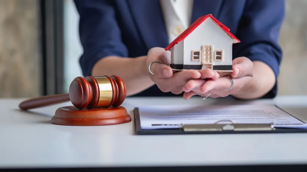 Person in blue blazer holding a small white house model with red roof near a wooden gavel and clipboard.