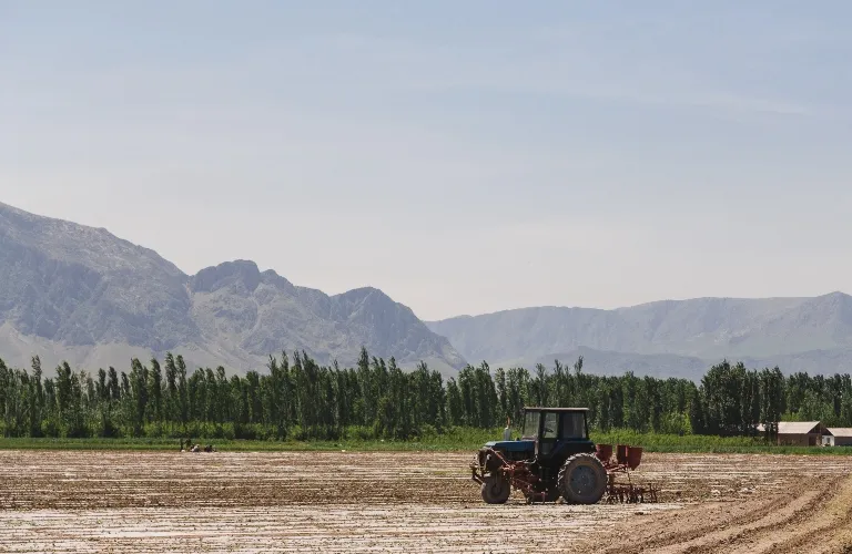 Blue tractor working on a plowed field with green trees and mountains in the background.