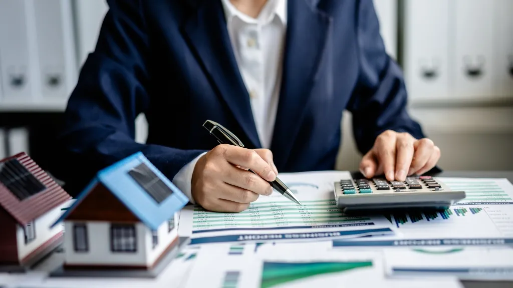 Professional in navy blazer using calculator and pen to analyze financial documents with miniature house models nearby.