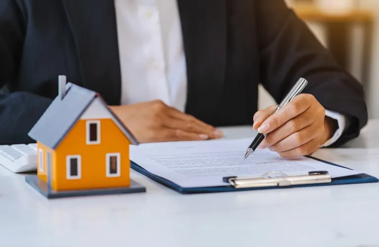 Person in formal attire signing a document beside a small orange house model on a white desk.