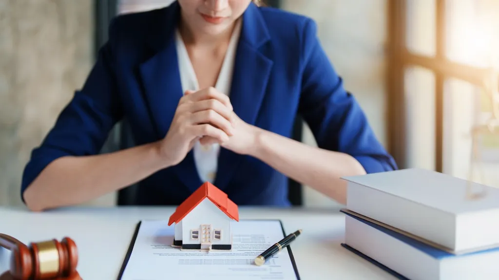 Professional woman in blue blazer sitting at desk with miniature house, legal documents, and gavel.